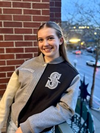 Photo of a teenage girl with long blonde hair. She is posing next to a brick wall and wearing a grey and black jacket with the letter S on it.