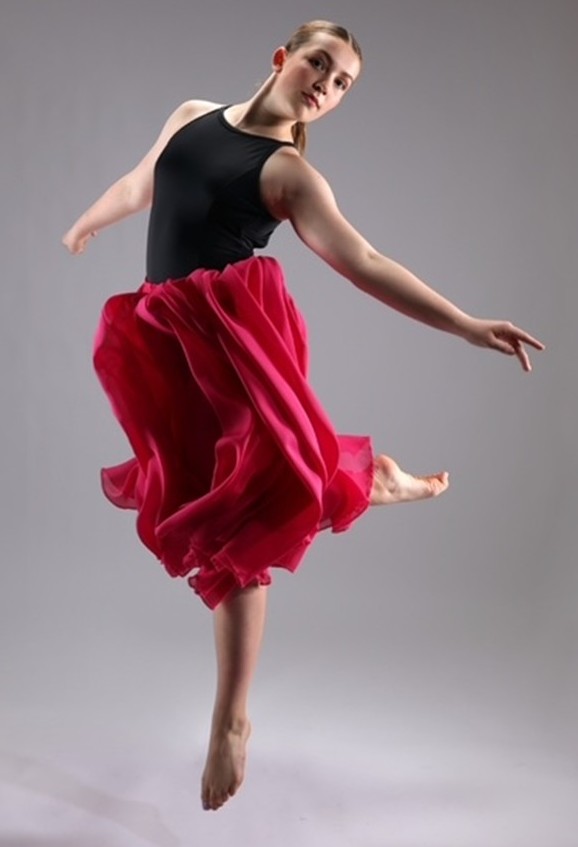 A teenage girl wearing a black leotard top and a billowing red skirt is executing a dance move in the air.