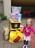 A little blonde girl wearing a light pink skirt and a bright pink hoodie is shown standing and smiling in front of many bins and bags of toys to donate to an annual toy drive.